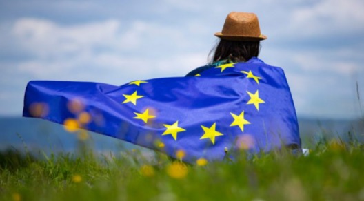 Immagine di una ragazza vista da dietro in un prato verde con bandiera Europa sulle spalle